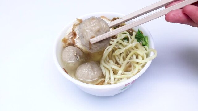 Indonesian culinary meatball soup containing noodles, tofu, celery and meat served in a white bowl on a wooden table. Eating meatballs with chopsticks, hands chopping meatballs