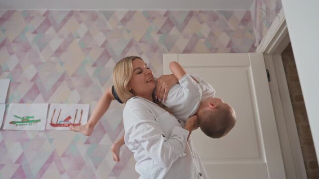 Playful mother embraces son happily. Laughing mother in white shirt lovingly lifts son in nursery. Cheerful family moment with mother swinging and hugging her son amidst vibrant decor