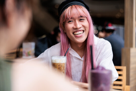 Person with pink hair smiles over drinks at cafe.