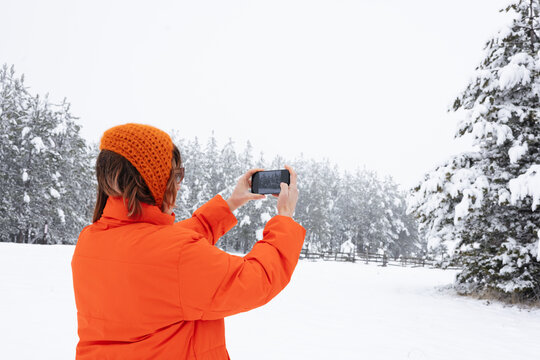 Woman In Orange Jacket Taking a Picture of Snowy Trees On Her Phone 