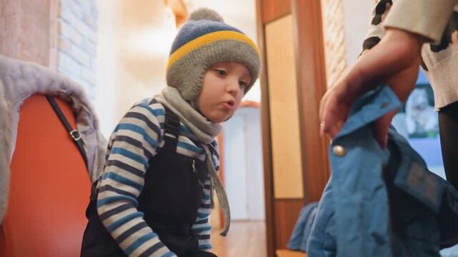 Young son sitting in hallway clutching toy, mom handing jacket, winter beanie and scarf, striped sweater, soft warm lighting, quiet anticipation of departure, gentle parental care