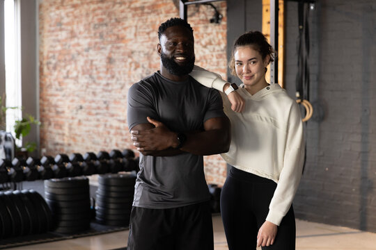 Diverse couple posing in fitness studio wearing workout gear and smartwatch with dumbbells, rings