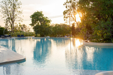 Obraz premium Luxury tropical resort swimming pool reflecting beautiful golden hour sunset light through green trees. Relaxing summer vacation, elegant hotel lifestyle, and peaceful nature travel getaway concept.