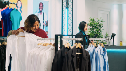 Obraz premium African american customer examining clothes on hangers and checking materials in clothing store. Young woman looking to buy casual or formal wear from shopping center, retail shop.