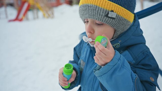 Little son blowing bubbles in snow at neighborhood playground, bundled in warm jacket and beanie, breath visible in cold air, joyful focus as fragile bubbles float, candid winter play moment full