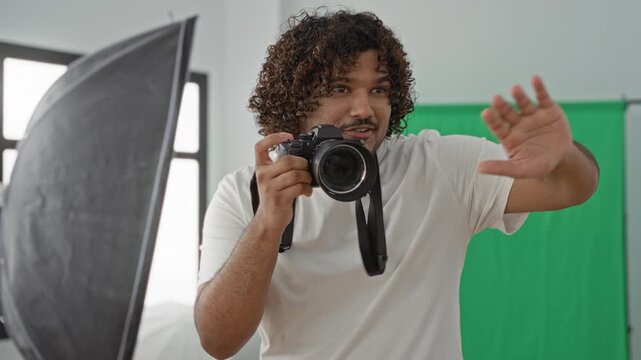 Man holds camera to eye, aiming and gesturing with hand to frame a shot in a studio with green backdrop and softbox; focused creativity.