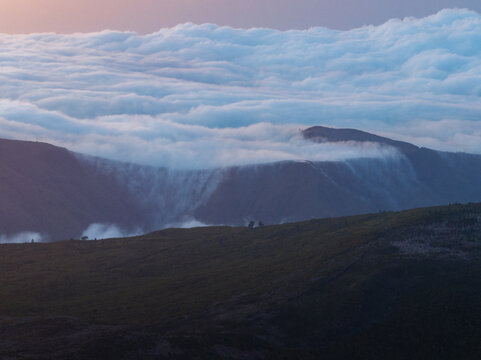 Aerial view of Bica da Cana with a thick sea of clouds flowing over the mountain ridges under a soft dawn sky in Ponta do Sol, Madeira, Portugal.