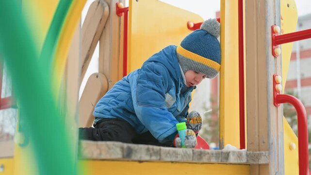 Son exploring playground bench with toy, mom nearby during winter outing. Lively movement as child inspects colorful toy, bundled jacket and pompom hat, snow traces on concrete, bright playground