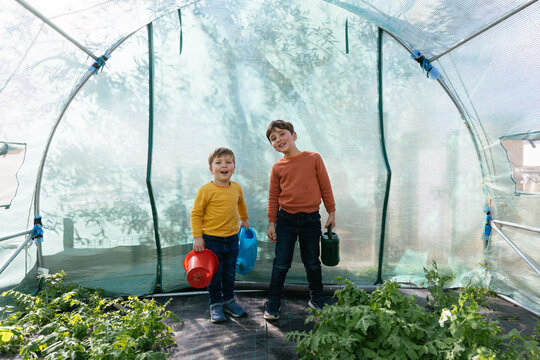 Children brothers smiling holding watering cans in greenhouse