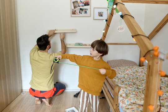 Father Installing Shelf While Son Measures in Bedroom