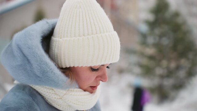 Winter mom adjusts hat, concerned gaze. Closeup portrait of protective mother in white knit beanie and scarf, blue coat, scanning snowy park for son. Visible breath in cold air, soft bokeh of trees