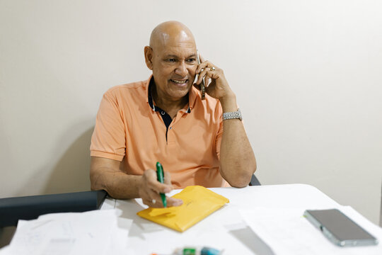 Man Talks on Phone While Taking Notes at Table in Home Office