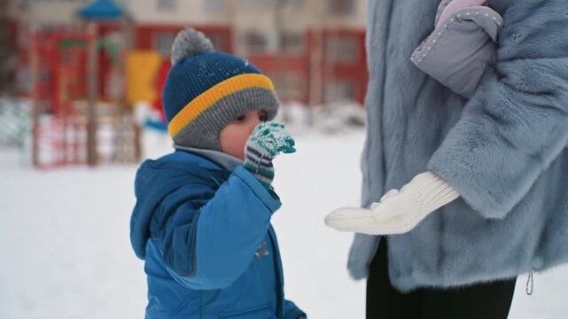 Snowy playground mom and son exchange cautious smile while mom in gray coat offers green shovel to boy in blue jacket and knit hat, winter urban backdrop, gentle guidance and family bond
