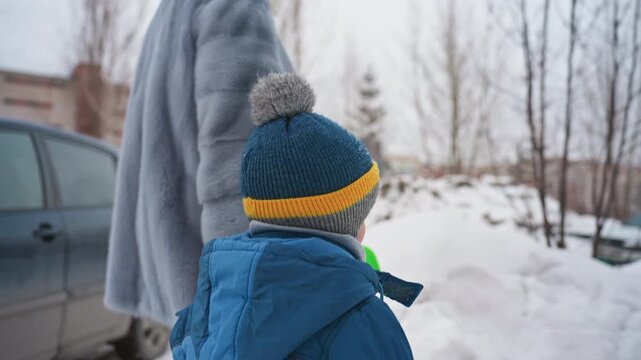 Winter parking lot scene mom and son, toddler in blue jacket and yellow trim beanie holds green shovel beside standing mother in grey coat near parked car and snow mound overcast sky, suburban