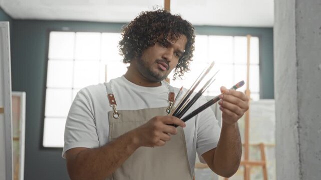 Man in apron inspects a fan of paintbrushes, turning and selecting bristles with bare hands while standing by easels and canvases in studio; concentration.