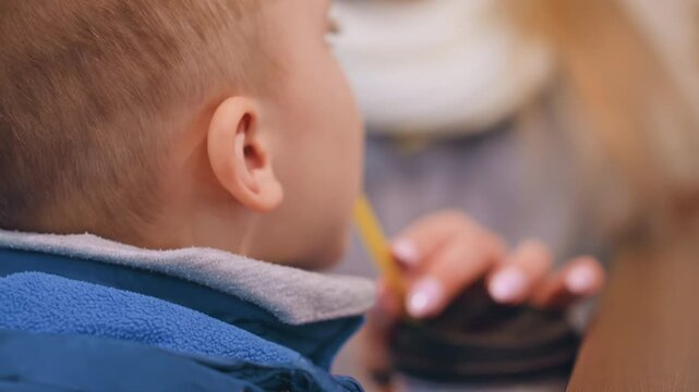 Mom and son sharing drink at cafe, mother offering cup with straw bluejacketed toddler sipping while mother smiles, white scarf and manicured nails visible, warm indoor ambience, tender connection