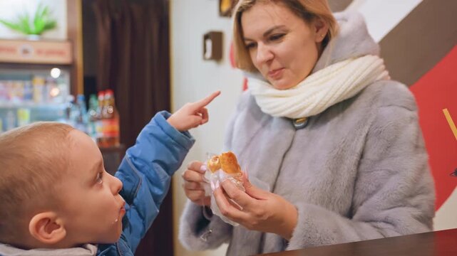 White mom feeding son pastry in cozy cafe, wearing scarf and coat, offering bite at counter next to coffee cup, warm lighting, candid family bond, gentle caregiver interaction, child enjoying snack