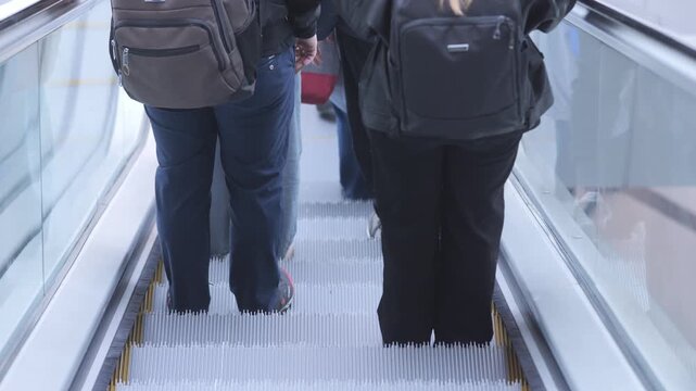 People ride escalator upward. Two individuals walk forward. One carries backpack. Another wears dark pants. Glass sides surround moving steps