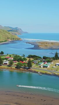 A boat speeds down the turquoise waters of the River in Marokopa, Waikato, New Zealand, passing by lush green hills and a small coastal town.
