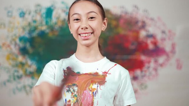 Young cute girl standing at colorful stained wall while holding brush and point at camera. Happy high school student wearing white shirt with stain color and look at camera with confident. Edification