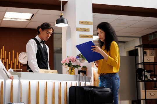 Asian guest signs registration form to stay at lavish and modern resort for holiday. Customer goes on vacation and arrives in hotel lobby with luggage, where concierge hands over necessary papers