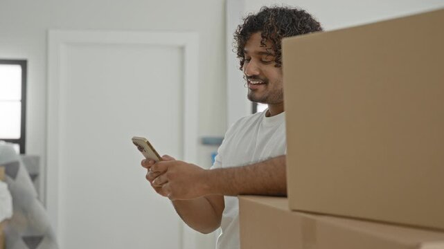 Man holds smartphone and smiles while leaning on stacked moving boxes in a building; moving day joy.