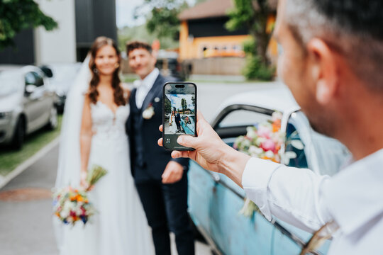 Guest photographing bride and groom beside vintage car at Lake M