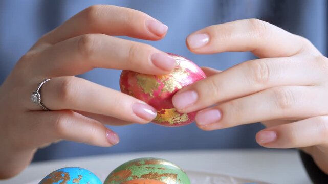 Close-up of hands holding a decorated red egg with gold patterns. Other painted eggs visible nearby. Fingers gently turn the egg for viewing. Background shows soft focus fabric