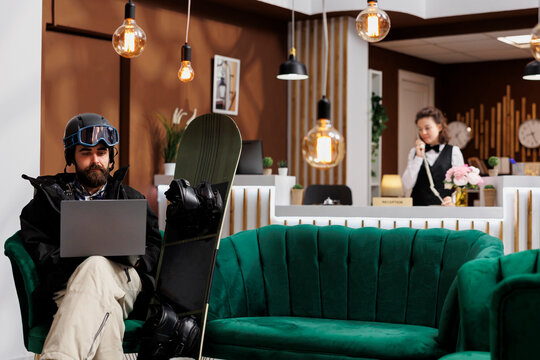 Male snowboarder sitting in hotel lobby utilizing internet on laptop for reservation checks. Caucasian man wearing in snow clothing using minicomputer in ski resort staff answers phone in background.