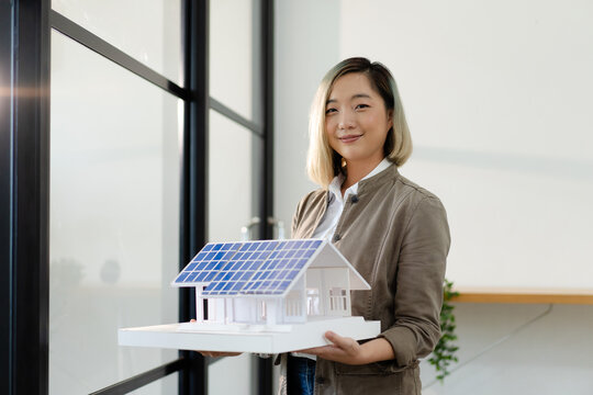 female architect engineer raising house model with solar panel standing near window, asian woman holding model smiling for new idea real estate property innovation while working at office