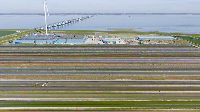 Aerial view of an industrial aquaculture facility with long rectangular ponds and a wind turbine near the Zeeland Bridge in the Netherlands.