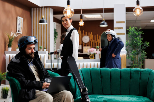 Caucasian man utilizing laptop to book activities for winter vacation at ski resort. Male guest dressed in snow clothing using minicomputer in lounge area while employee takes suitcase to hotel room.