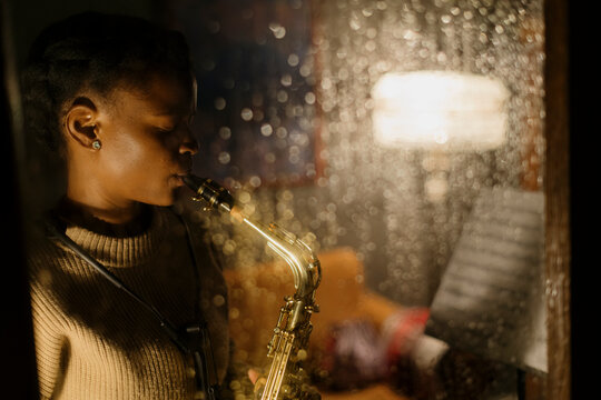Woman playing saxophone on rainy evening, viewed through window