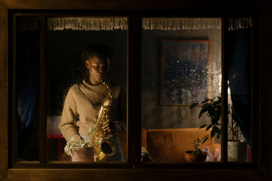 Woman playing saxophone on rainy evening, viewed through window