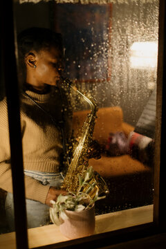 Woman playing saxophone on rainy evening, viewed through window