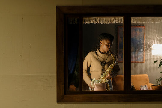 Woman playing saxophone on rainy evening, viewed through window