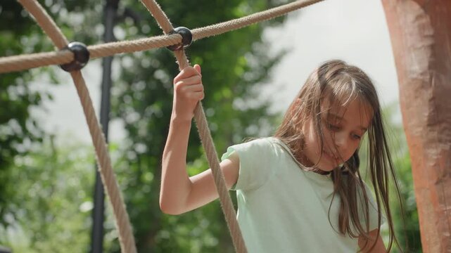 Young daughter climbing rope course in park, careful hands and braided hair, balancing between wooden posts under bright sky, focused ascent and playful determination, outdoor adventure building