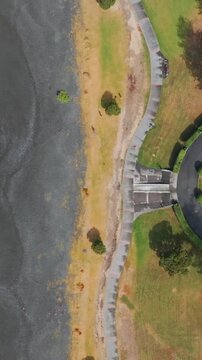 Aerial view of the beach and a circular fountain at Marine Parade, Napier, Hawkes Bay, New Zealand. The turquoise ocean meets the dark sand beach, with a park and street visible.