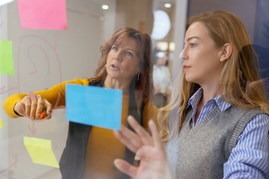 Businesswomen brainstorming ideas on glass wall with sticky notes