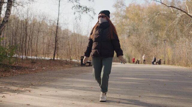 Female walking near wooded park edge, glancing sideways while pacing along paved path, wearing warm jacket, beanie and backpack with face mask, scattered leaves and soft sunlight, calm vigilant mood