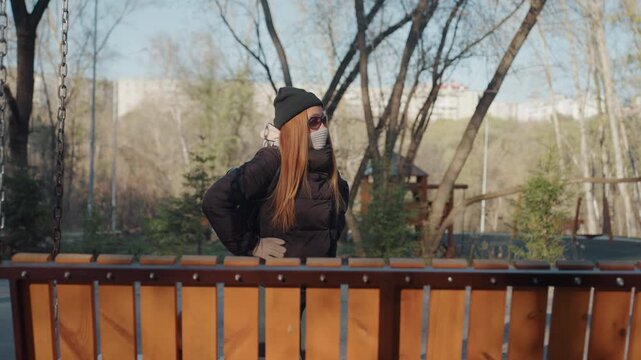 Park caucasian woman hands on hips defiant stance, masked redhead in beanie and sunglasses at wooden railing, winter jacket and confident gaze, watchful posture suggests suspected snatcher or vigilant
