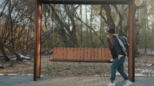 Cautious approach near wooden bench, lone individual with backpack pauses and scans surroundings like a prowler, tense winter light, leaves and sparse snow, scene suggests attempted snatch and quick