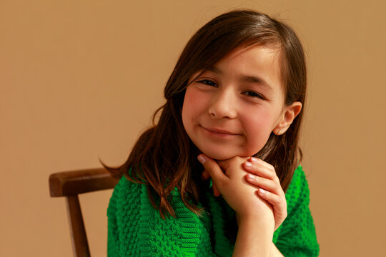 A Young Girl Sits on a Wooden Chair With Her Hands Under Her Chin