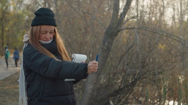 White woman pointing pen while recording outdoors, beanie and mask under chin, notepad held steady by river path, brisk autumn light and bare branches, determined reporter energy and documentary