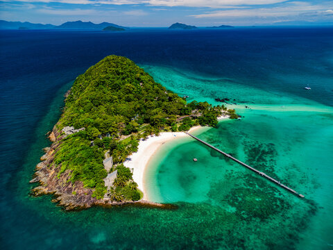 Aerial view of Ko Kham island featuring a lush green hill, white sand beach, and a long wooden pier stretching into turquoise coral reefs in Ko Mak, Trat, Thailand.