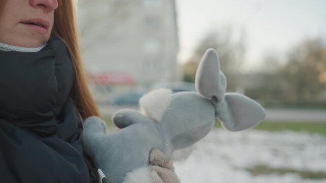White woman adjusts headphones holding gray bunny in chilly park, cap and coat visible, mittened grip on plush, wary glance and steady posture, urban sidewalk and light snow creating cautious mood