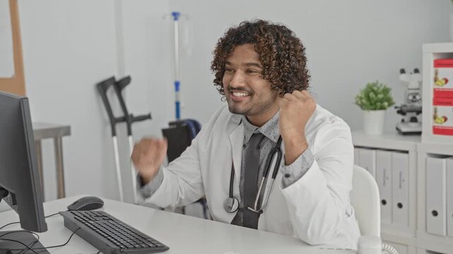 Man doctor in white labcoat with stethoscope raising fists and clapping at computer workstation in building, smiling broadly; success joy confidence.