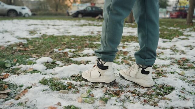 Closeup feet walking on snow, lone figure in jeans and white sneakers treads across icy park grass leaving crisp footprints cautious, stealthy steps with sole tread visible, ambient winter light,