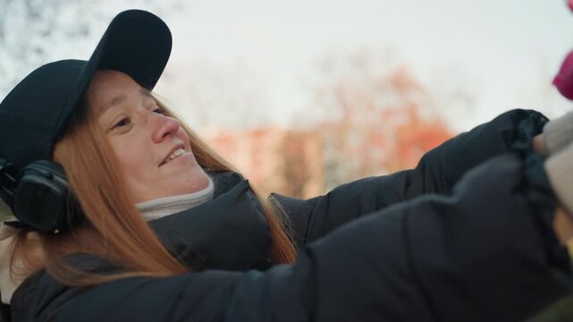 Reaching white woman grabs stuffed rabbit in park gloved hands stretch forward, playful snatcher vibe, black cap and headphones, puffer jacket and backpack, candid exchange with friend, autumn trees