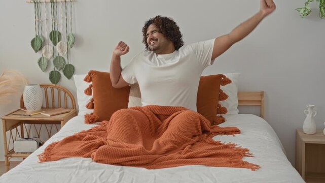 Young indian man on bed stretches arms up, yawning with orange blanket, tassel pillow and wicker bedside table in studio; morning serenity.